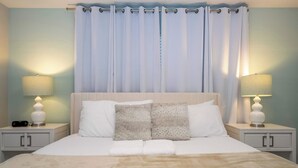 Ein ordentlich bezogenes Bett mit weißen Leinen und einer beigen Tagesdecke, begleitet von zwei Nachttischen mit Lampen und einer Digitaluhr.