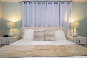 Cama bem arrumada com lençóis brancos e manta bege, com duas mesas de cabeceira de cada lado, com luminárias e um relógio digital.