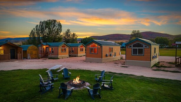 A row of cabins with a fire pit and chairs in the foreground, set against a backdrop of hills and a sunset sky.