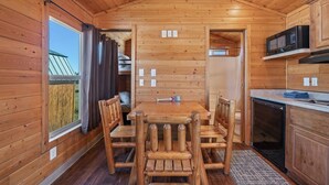 A wooden interior with a dining table and chairs, a kitchen area with a microwave, and a view of the outside. - #K305 Quail Cabin - AC, Fire Pit & BBQ! (Saint Charles)
