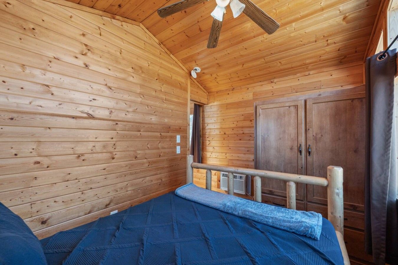 Una habitación de cabaña de madera con una cama, un ventilador de techo y una puerta que da a otra habitación.