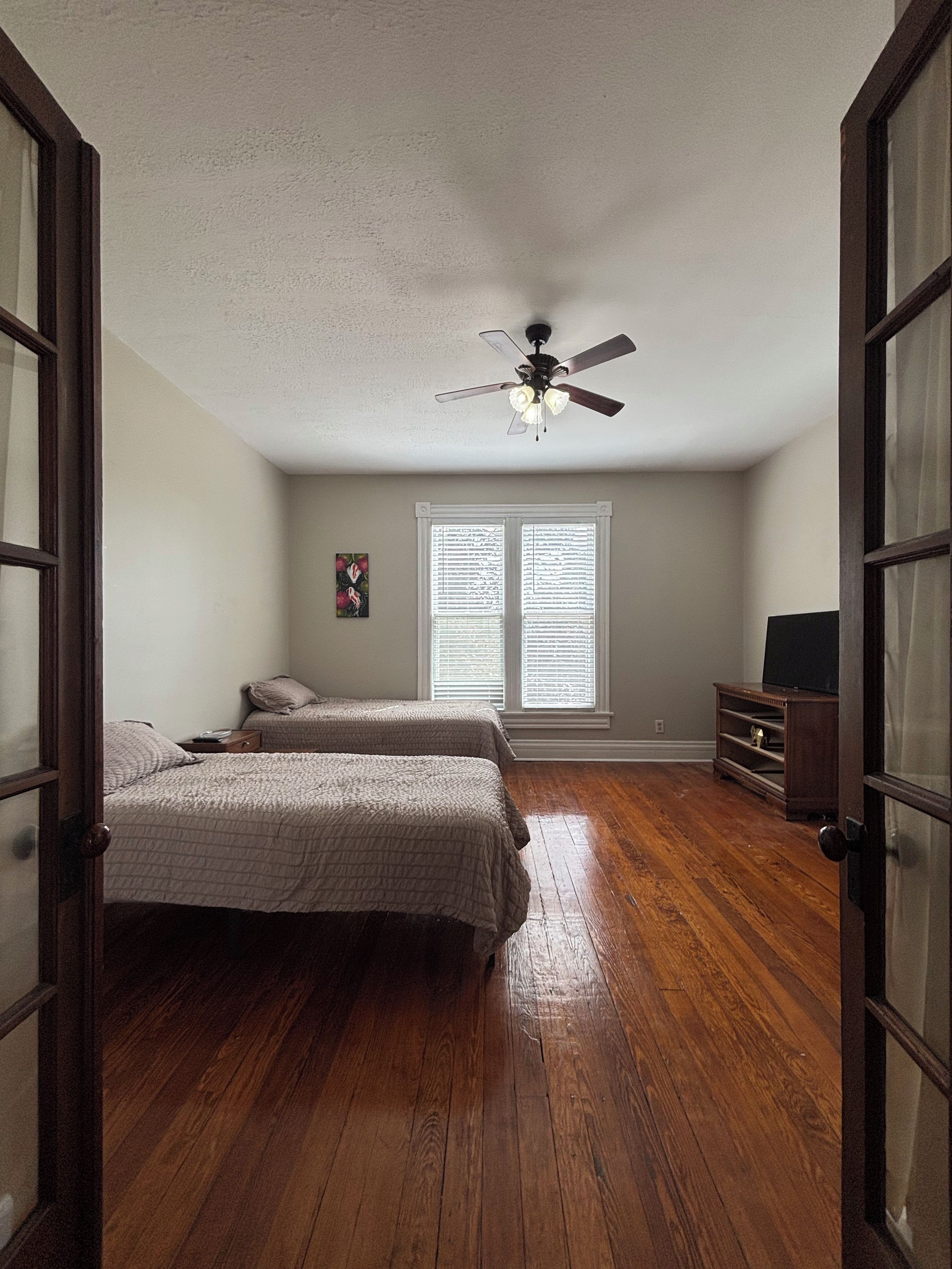 Une chambre avec deux lits, un ventilateur de plafond, une fenêtre avec des stores et un téléviseur posé sur un meuble en bois.