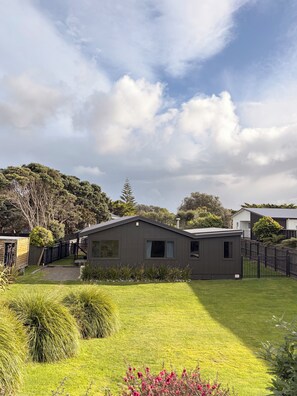 A residential property with a well-maintained lawn, a fenced yard, and a house with a dark exterior. - Kiwiana themed bach, perfect for family getaways or a retreat near the beach! (Waikanae Beach)