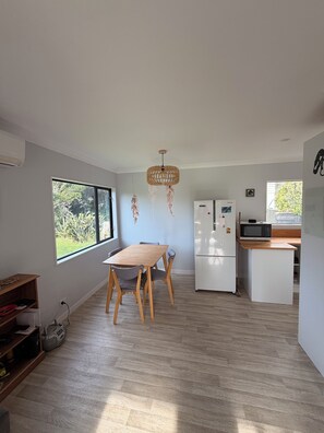 A modern kitchen with a dining table, white appliances, and a large window. - Kiwiana themed bach, perfect for family getaways or a retreat near the beach! (Waikanae Beach)