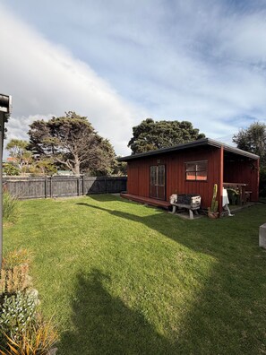 A red wooden building with a porch, surrounded by a well-maintained lawn and a wooden fence. - Kiwiana themed bach, perfect for family getaways or a retreat near the beach! (Waikanae Beach)