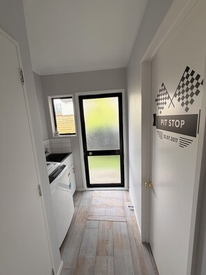 A narrow hallway with a kitchen area on the left, a large frosted glass door, and a door with 'PIT STOP' signage. - Kiwiana themed bach, perfect for family getaways or a retreat near the beach! (Waikanae Beach)