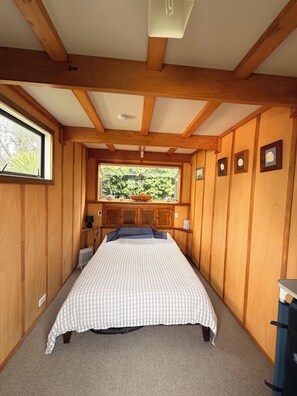 A bedroom with a wooden ceiling, a large window with a view of greenery, a bed with a patterned bedspread, and a radiator. - Kiwiana themed bach, perfect for family getaways or a retreat near the beach! (Waikanae Beach)