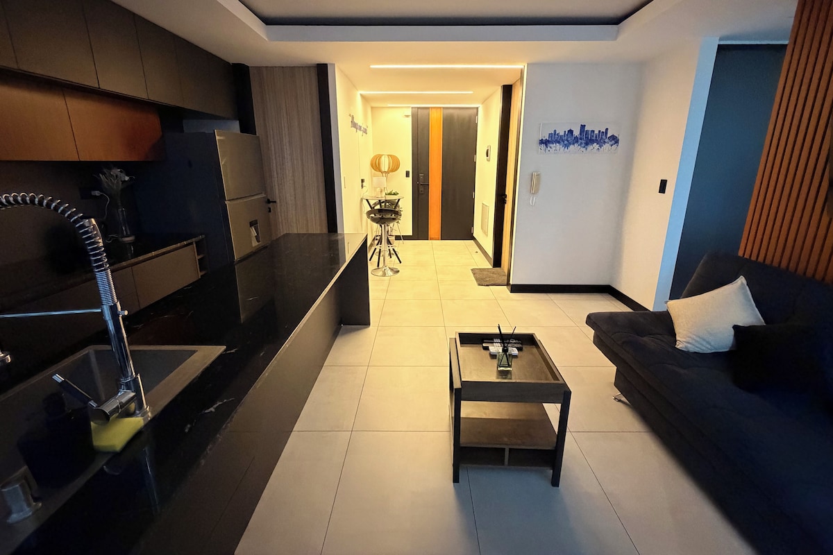 Una sala de estar moderna con cocina integrada, un sofá, una mesa de centro y una mesa de comedor con sillas.