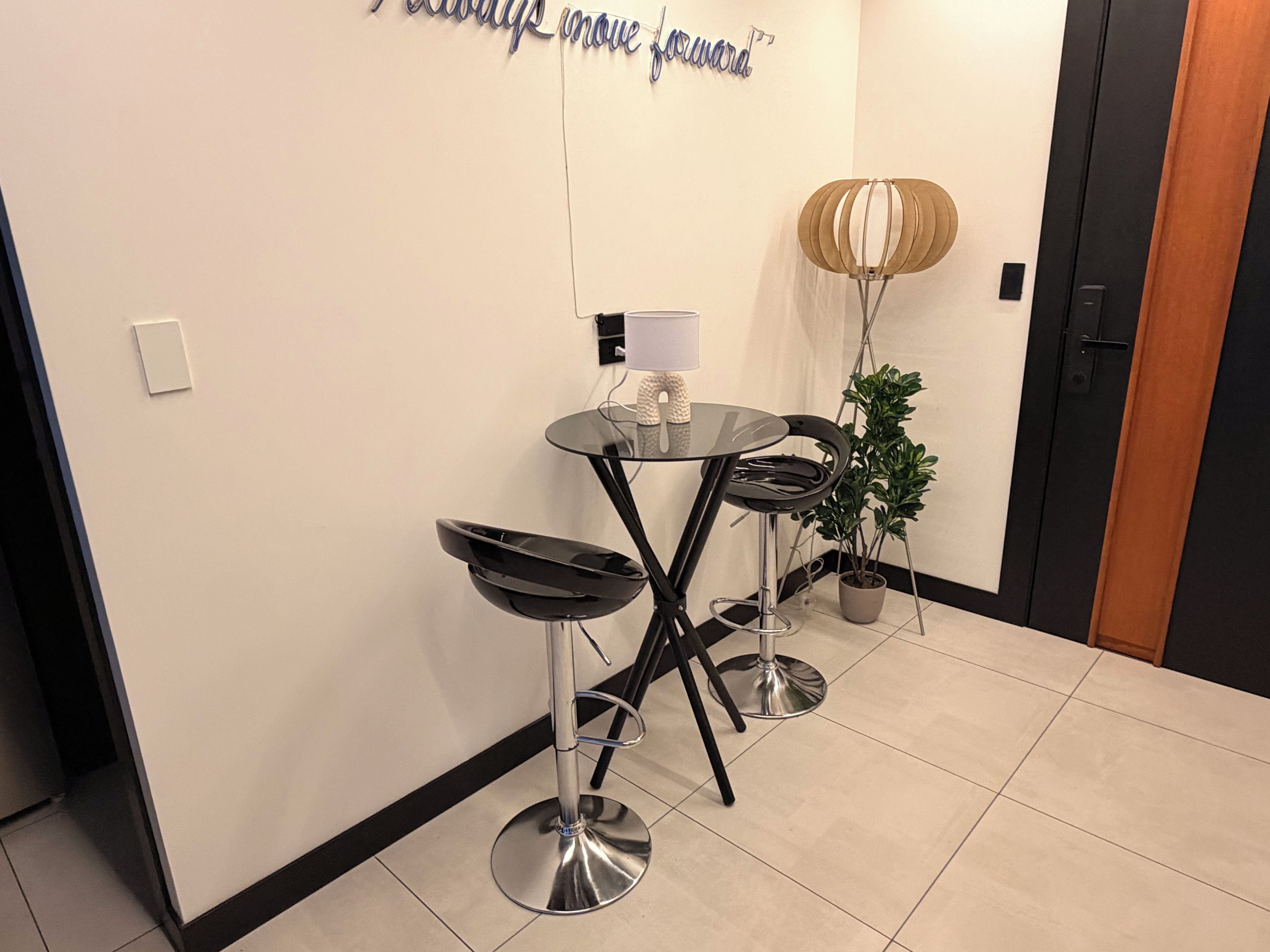 Ein modernes Interieur mit einem Glastisch, zwei schwarzen Barhocker, einer Topfpflanze und einer Wandlampe.