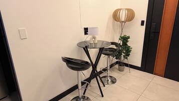 Ein modernes Interieur mit einem Glastisch, zwei schwarzen Barhocker, einer Topfpflanze und einer Wandlampe.
