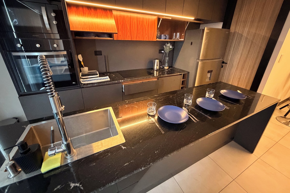 A modern kitchen with a black countertop, stainless steel appliances, and a wooden backsplash.