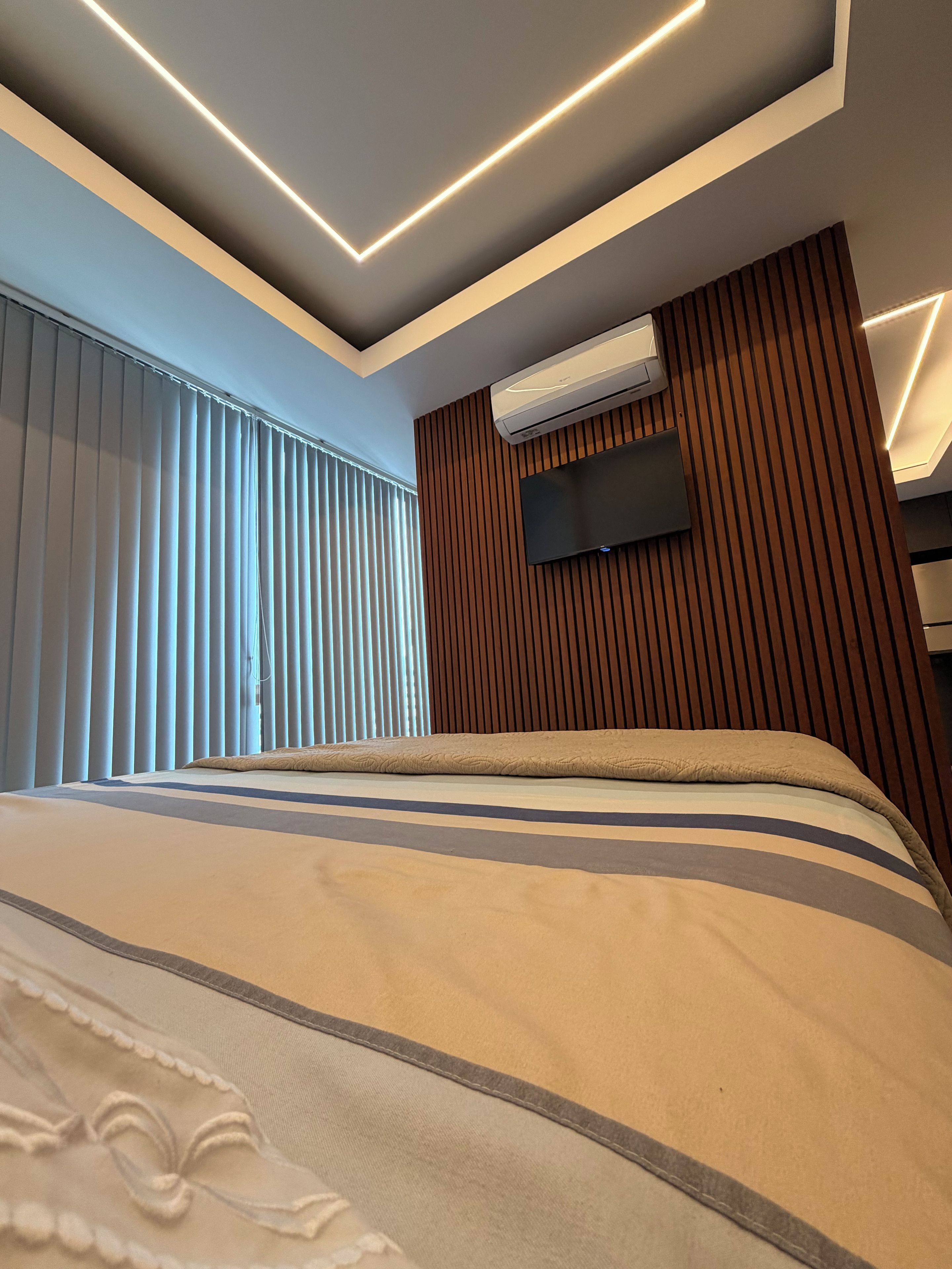 A modern bedroom with a large bed, a flat-screen TV mounted on a wooden panel wall, and an air conditioning unit.