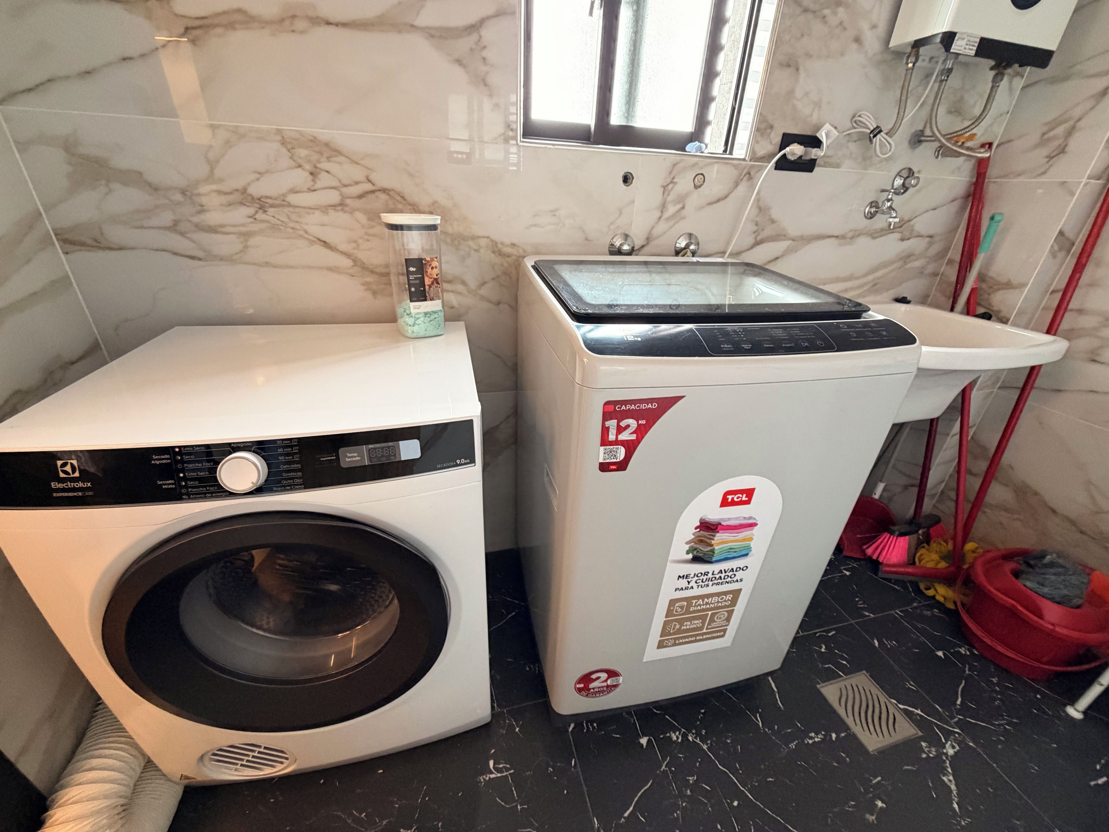 Eine kompakte Wasch- und Trocknerkombination mit einer Kapazität von 12 kg, in einem Badezimmer mit Marmorboden und einem Fenster oberhalb der Einheit.