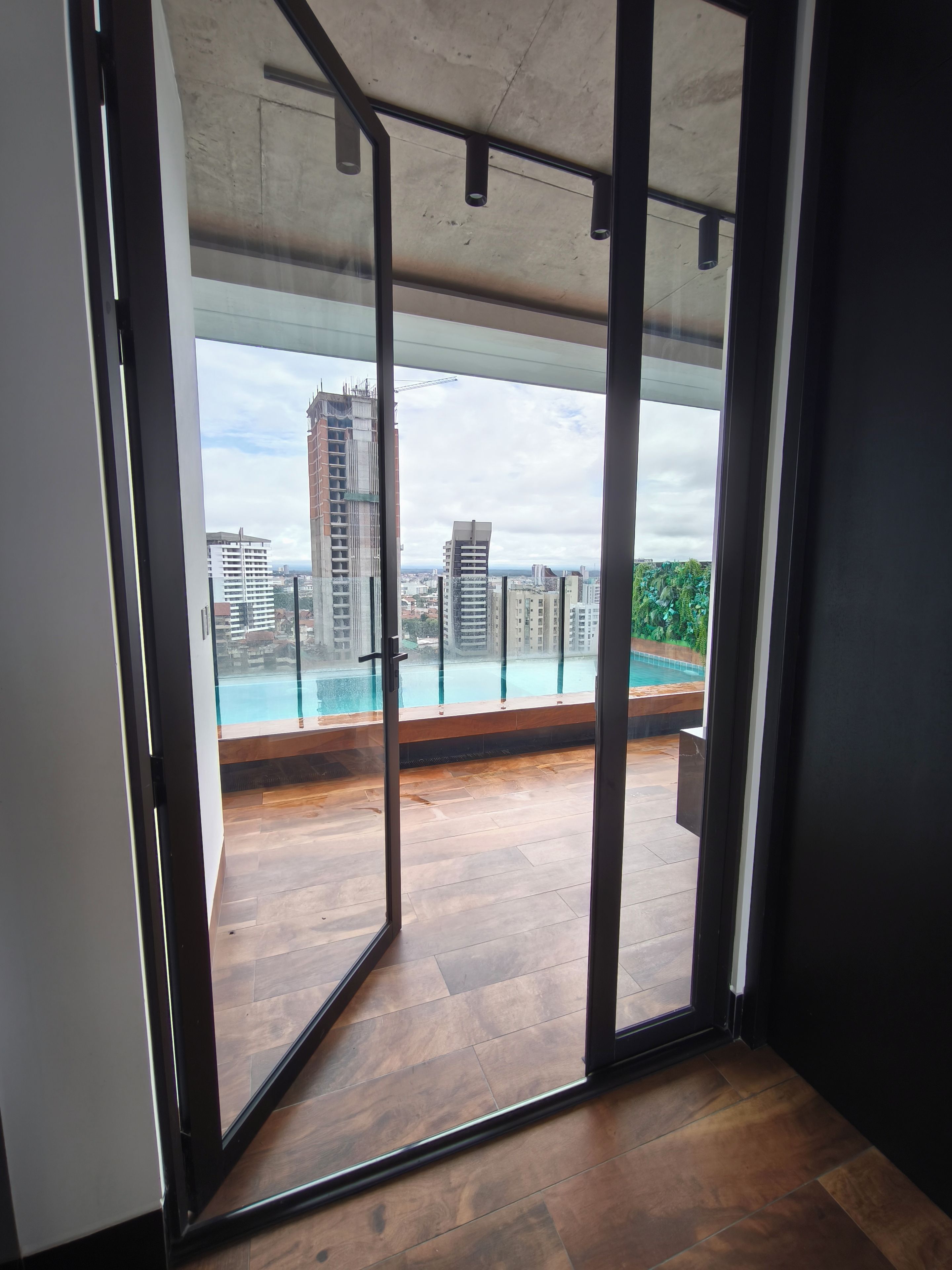 A modern interior with a large glass door leading to an outdoor balcony with a view of a swimming pool and surrounding buildings.