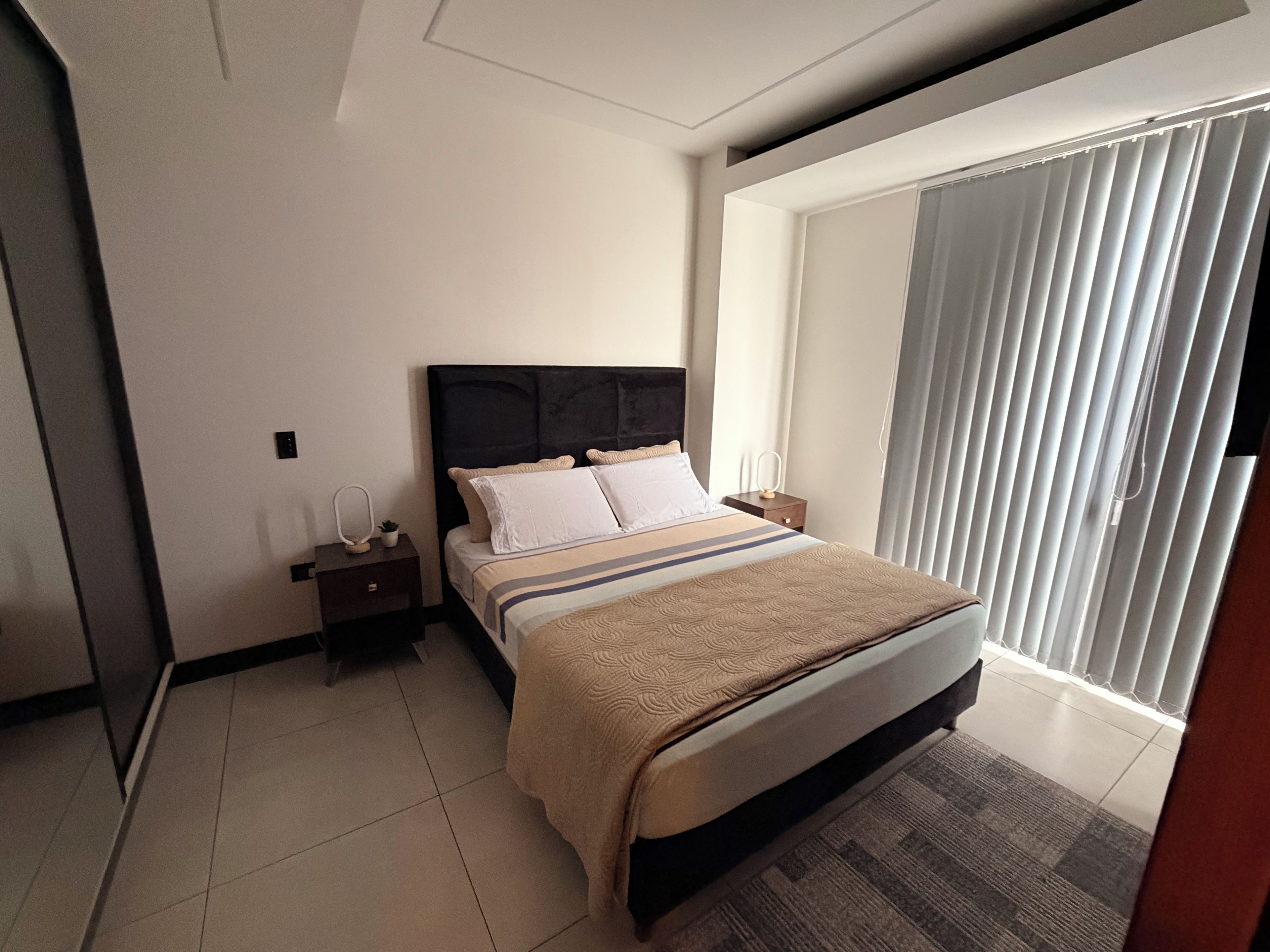 Habitación de hotel con cama, mesita de noche, lámpara y un ventanal grande con persianas.