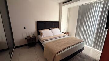 Habitación de hotel con cama, mesita de noche, lámpara y un ventanal grande con persianas.