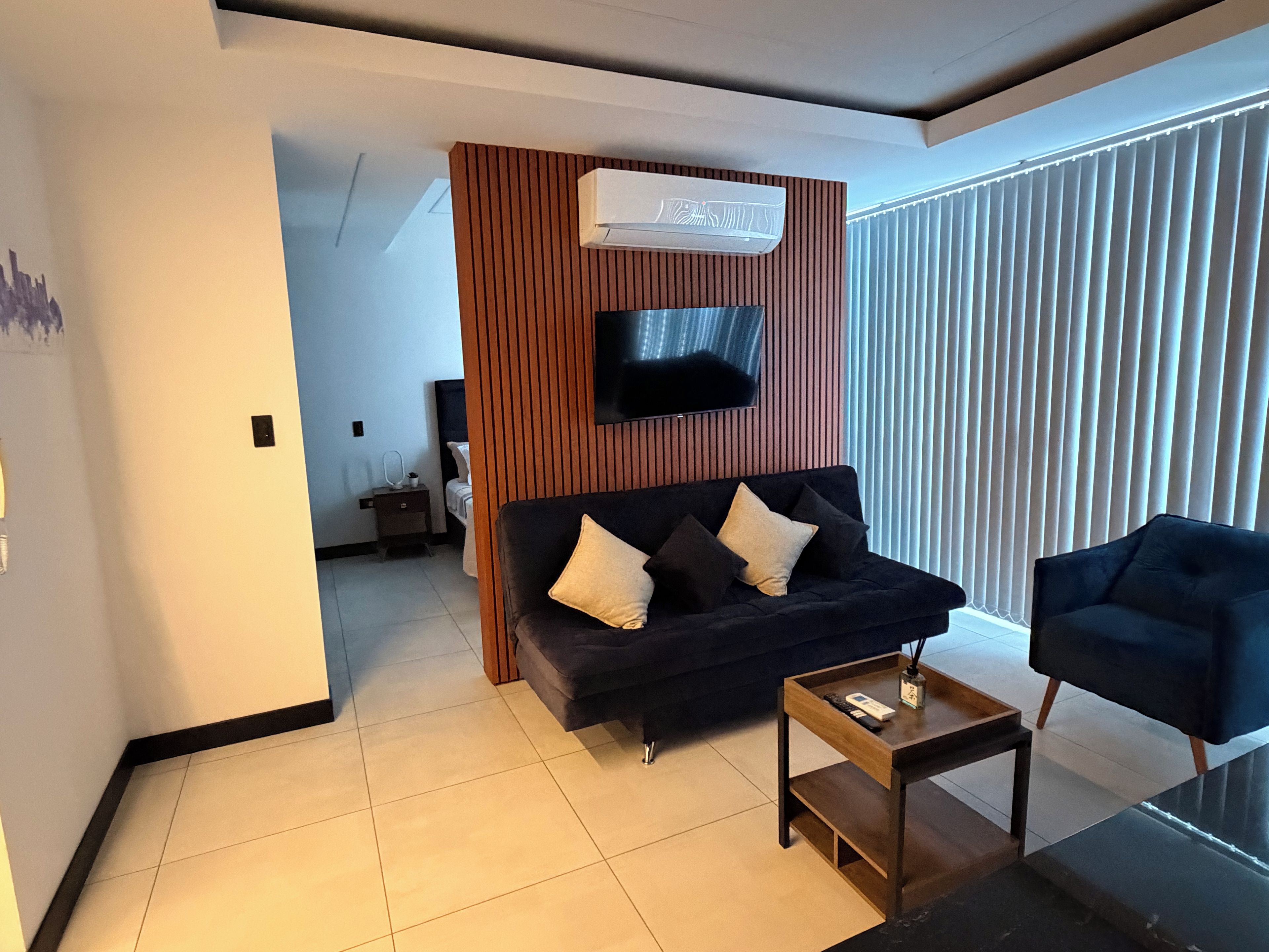 Una sala moderna con un sofá oscuro, una pared de acento de madera, un televisor de pantalla plana y un ventanal con persianas.
