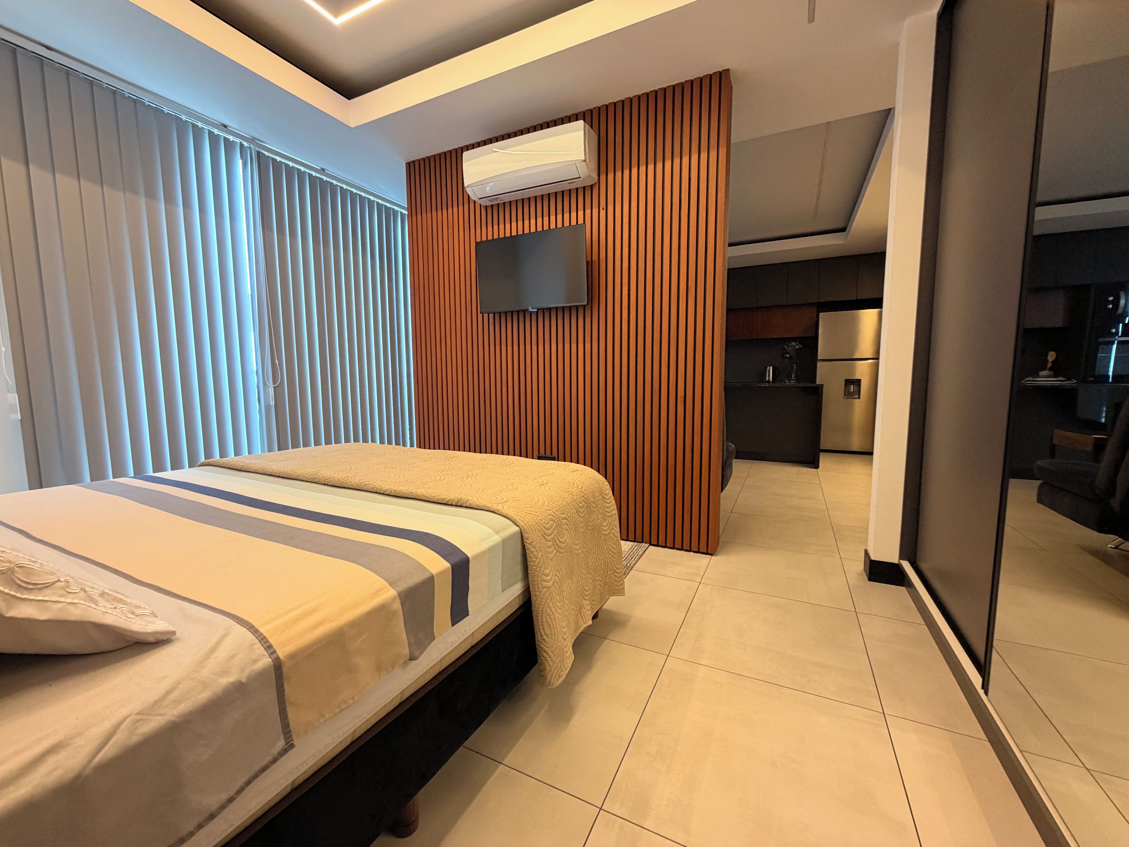 Une chambre d’hôtel moderne avec un lit, un téléviseur fixé au mur, un mur recouvert de panneaux en bois et, au fond, une petite cuisine.
