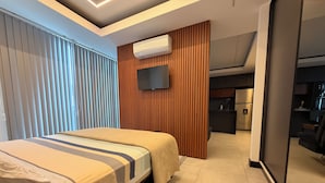 A modern hotel room with a bed, a TV mounted on the wall, a wooden panel wall, and a kitchenette in the background. - 518 RENT (Santa Cruz)