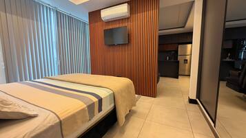 Une chambre d’hôtel moderne avec un lit, un téléviseur fixé au mur, un mur recouvert de panneaux en bois et, au fond, une petite cuisine.