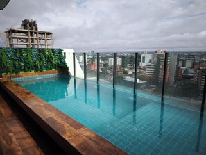 Piscina infinita en la azotea con vista a la ciudad, rodeada por una terraza de madera y un muro verde con plantas.