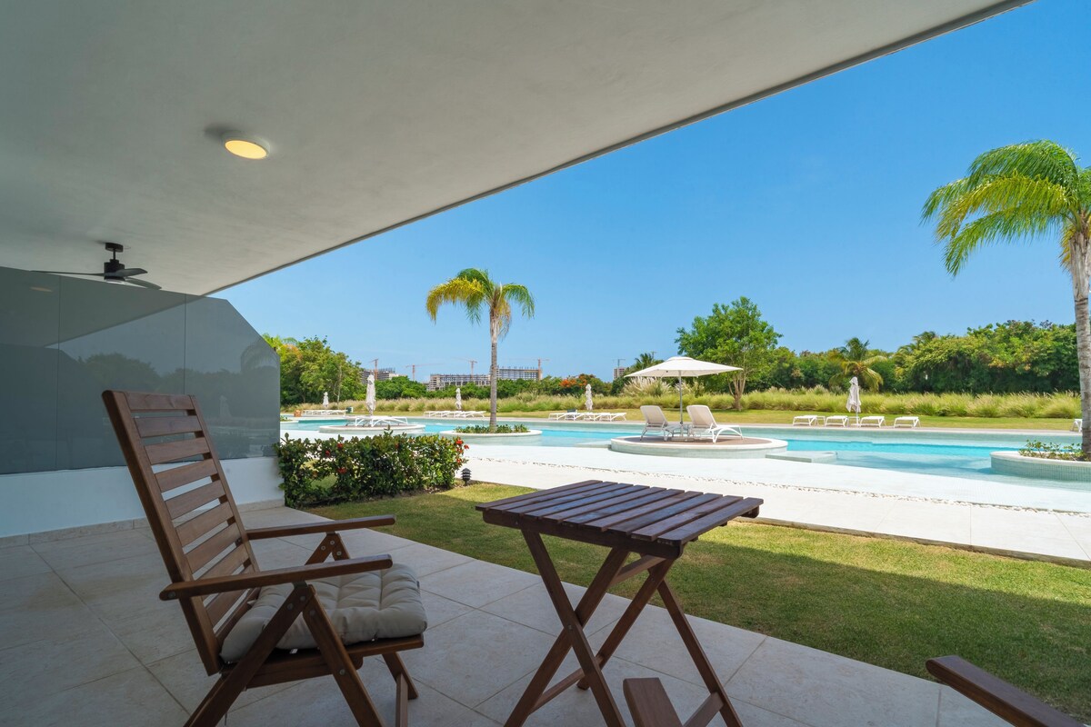 Zona junto a la piscina con una silla de madera, una mesita y vistas a una piscina y palmeras.
