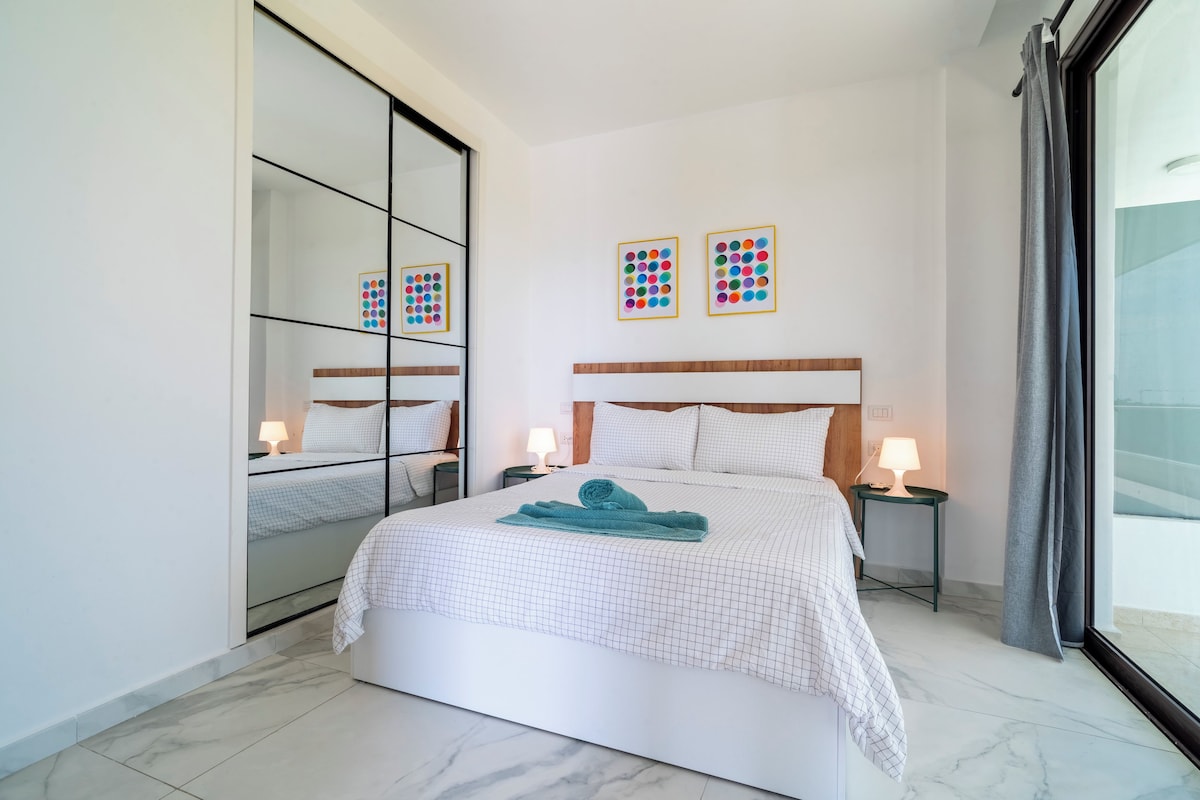 Un dormitorio moderno con una cama grande, dos mesitas de noche con lámparas, un espejo y dos cuadros en la pared.