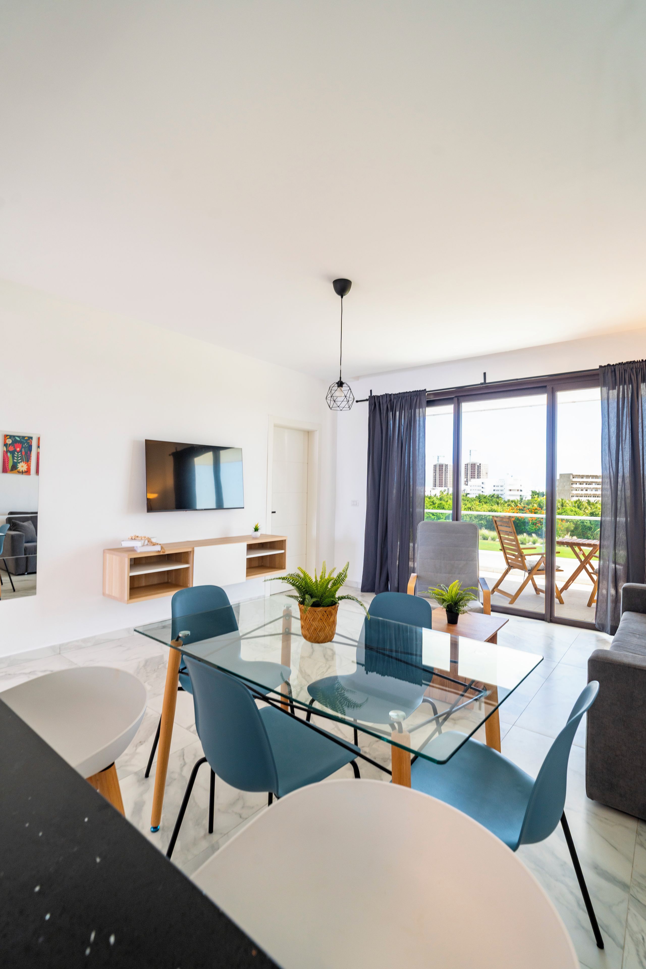 Un salon moderne avec une table à manger en verre, des chaises bleues, un canapé et un téléviseur fixé au mur.