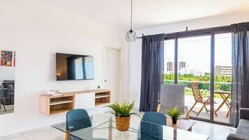 A modern living room with a glass dining table, blue chairs, a sofa, and a television on the wall.