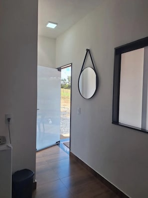 Un couloir avec une porte blanche donnant sur l’extérieur, un miroir rond accroché au mur et une fenêtre à cadre noir.