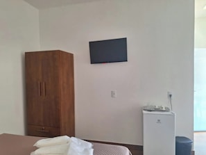 A room with a bed, a wooden wardrobe, a wall-mounted television, and a small white appliance.