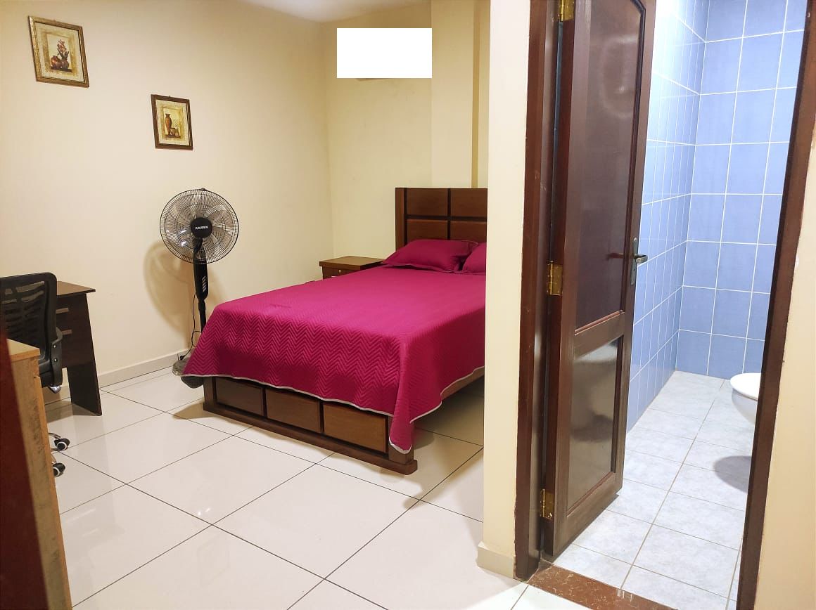 Une chambre à coucher avec un lit, un bureau, une chaise, un ventilateur sur pied, et une salle de bain visible à travers une porte ouverte.
