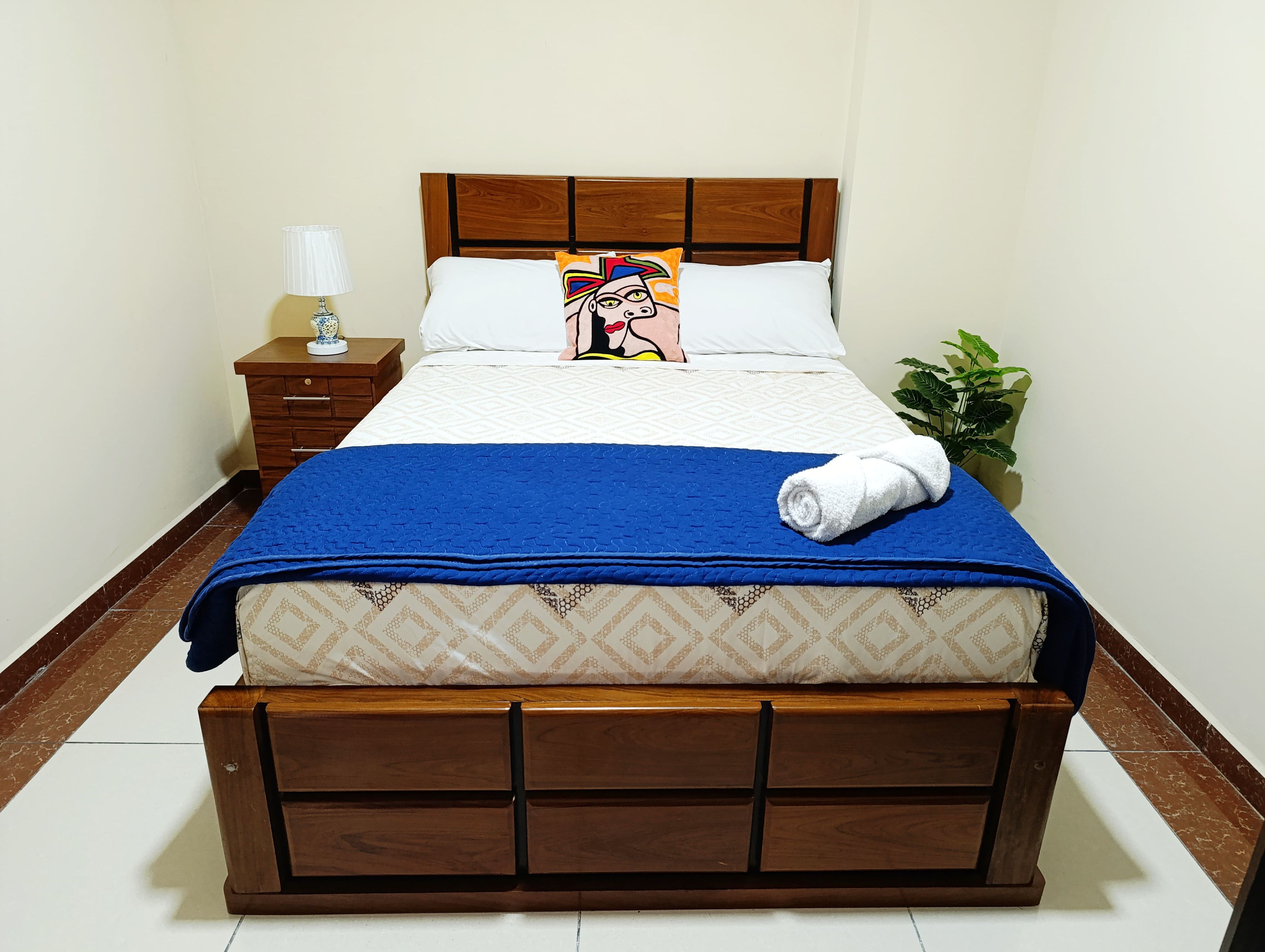 A neatly made bed with a blue blanket, a pillow with a colorful design, and a white towel.