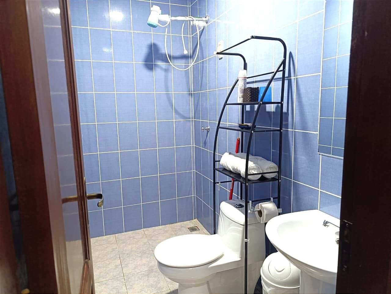 Un baño con paredes de azulejos azules, inodoro y lavamanos blancos, y una repisa metálica con toallas.