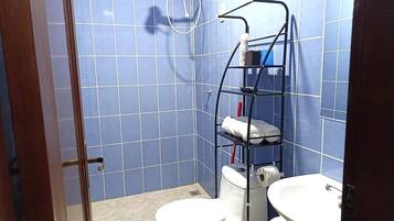 Un baño con paredes de azulejos azules, inodoro y lavamanos blancos, y una repisa metálica con toallas.