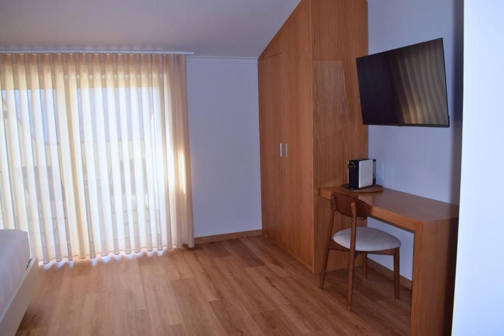 A room with a wooden floor, a desk with a chair, a television mounted on the wall, and a window with vertical blinds.