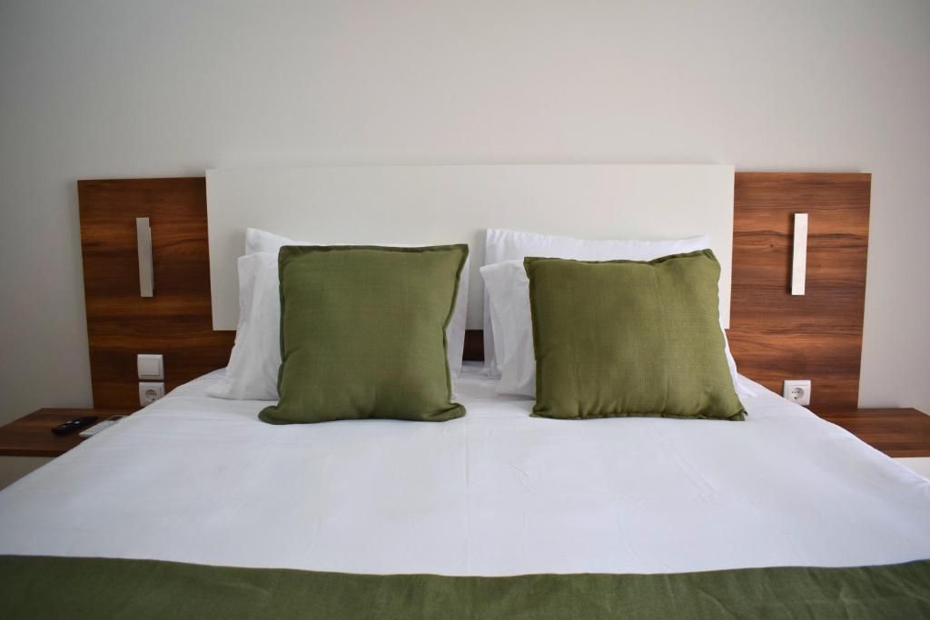 A neatly made bed with white linens and two green pillows, a wooden headboard, and a bedside table with a remote control.