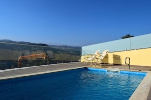 Una piscina con muelle de madera, sillas de descanso y barandilla de vidrio.