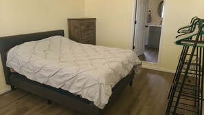 Een slaapkamer met een bed, een houten dressoir, een zichtbare badkamer door een geopende deur en een metalen rek met kleding.