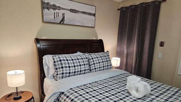 Una habitació d'hotel amb un llit, un capçal, una taula de nit amb una làmpada, una finestra amb cortines i un quadre penjat a la paret.
