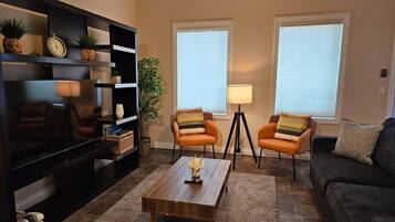 Un salon équipé d’un téléviseur à écran plat, de deux fauteuils, d’une table basse et d’un canapé. Il y a deux fenêtres avec des stores, une lampe et une bibliothèque agrémentée d’objets décoratifs.