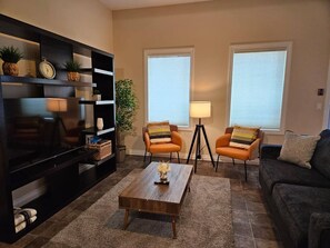 Ein Wohnzimmer mit Flachbildfernseher, zwei Sesseln, einem Couchtisch und einer Couch. Zwei Fenster mit Jalousien, eine Lampe und ein Bücherregal mit Dekorationselementen sind ebenfalls vorhanden.