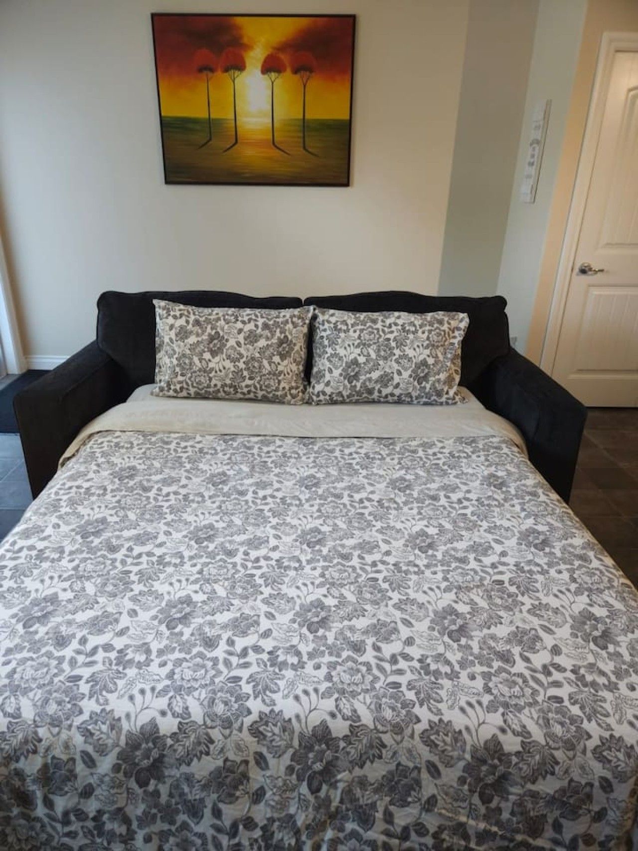A bed with a floral-patterned bedspread, two matching pillows, and a painting of trees on the wall.
