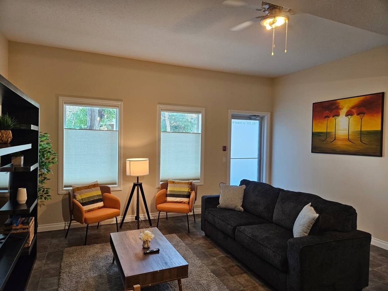 A living room with a sofa, two armchairs, a coffee table, and a lamp. There are windows with blinds, a painting on the wall, and a ceiling fan.
