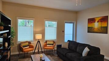A living room with a sofa, two armchairs, a coffee table, and a lamp. There are windows with blinds, a painting on the wall, and a ceiling fan.