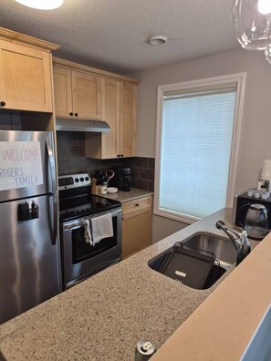A modern kitchen with stainless steel appliances, wooden cabinets, a window with blinds, and a welcome sign.