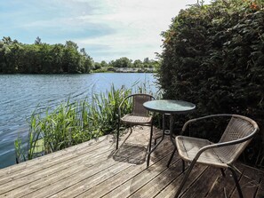 A wooden deck with two wicker chairs and a small round table, overlooking a calm body of water with a row of houses in the background. - Tattershall Lakes Country Park - Holiday Accommodation 16408 (Lincoln)