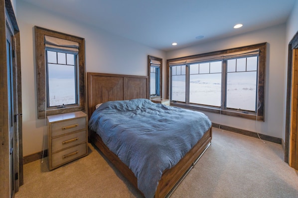A bedroom with a large bed, wooden headboard, a nightstand, and a dresser. There are two large windows providing a view of a snowy landscape.