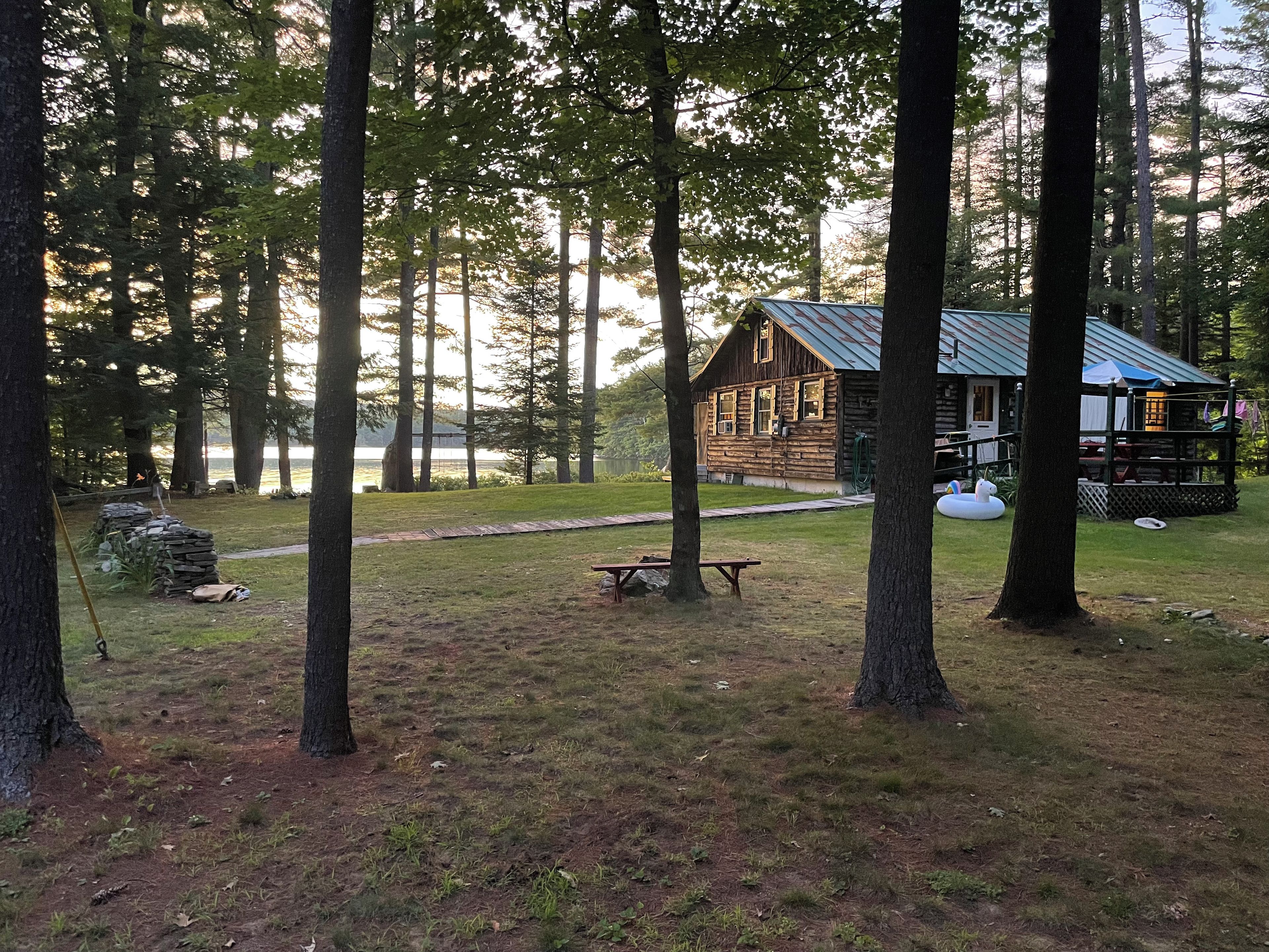 A cabin with a deck, surrounded by trees and a grassy area with a picnic table.