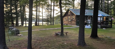 A cabin with a deck, surrounded by trees and a grassy area with a picnic table.